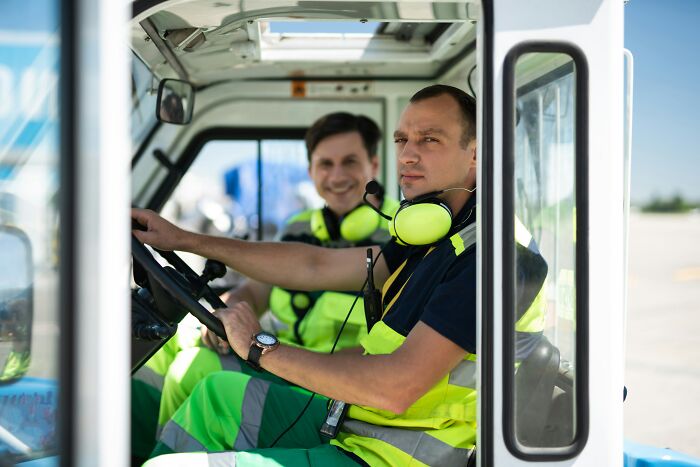 Two men in high-visibility vests and headphones sitting inside a vehicle, representing suspicious professions together.