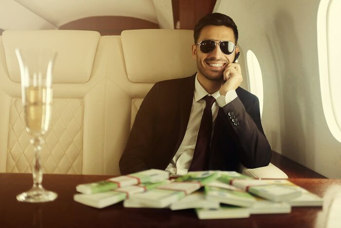 Confident man in suit and sunglasses smiling on phone inside private jet with stacks of money on table, representing success.