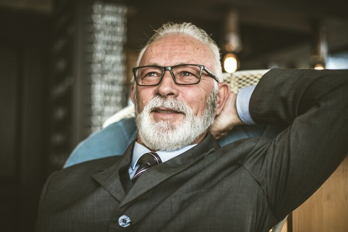 Elderly man with glasses and white beard relaxing in a suit, representing servers overhearing crazy stories.