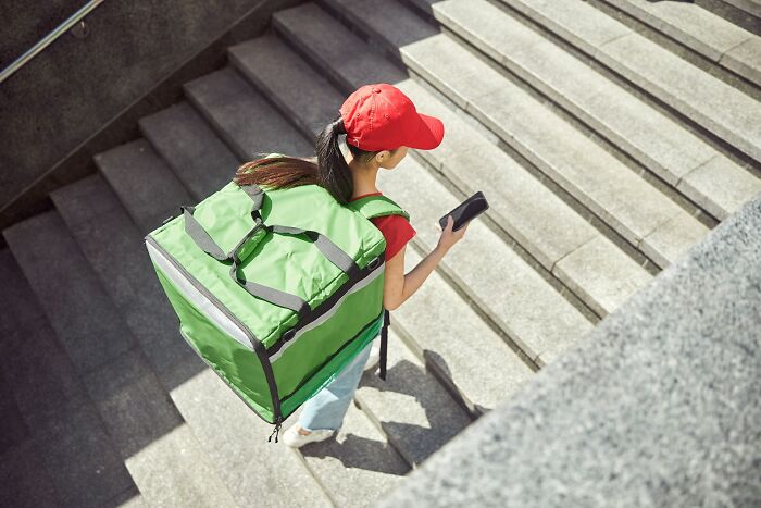 Delivery person wearing red cap and large green backpack walking down stairs while checking phone, travel complaints concept.