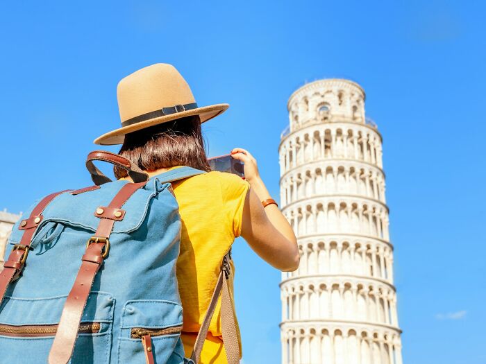 Traveler with a backpack taking a photo of the Leaning Tower of Pisa, one of the famous travel destinations seen in person.