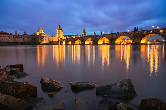 Illuminated historic bridge over calm river at dusk, showcasing real life scenes more interesting and bizarre than fiction.