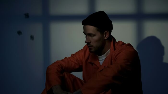 A man in an orange jumpsuit and black beanie sitting alone in a dimly lit room, reflecting on school bullies.