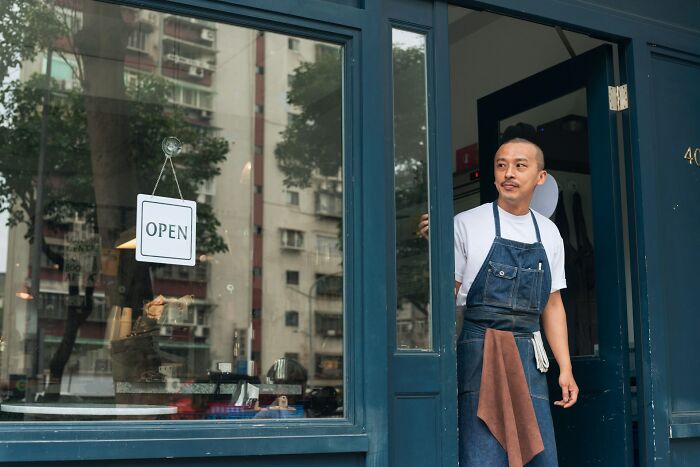 Server standing at restaurant entrance wearing apron with open sign on door, illustrating crazy stories servers overheard.