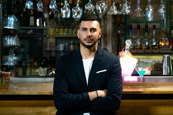 Man in a blazer standing with arms crossed behind a bar, representing professions not seen as relationship material.