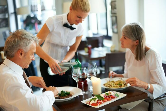 Waitress serving wine to a couple dining in a restaurant, overhearing crazy stories servers share with guests.