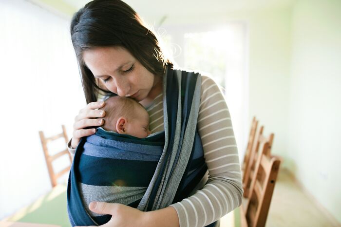 Adult woman gently holding a baby in a sling indoors, illustrating moments of caring and delayed growing up.