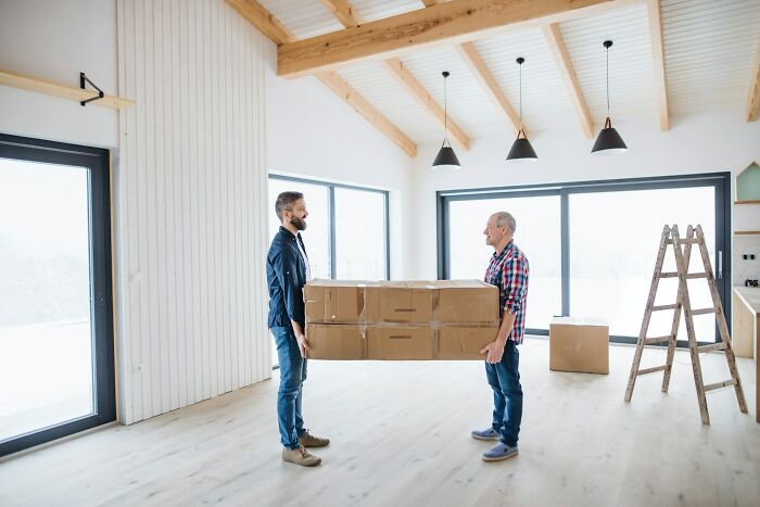 Two men carrying large cardboard boxes in a bright, modern home with wooden beams and large windows, moving day scene.