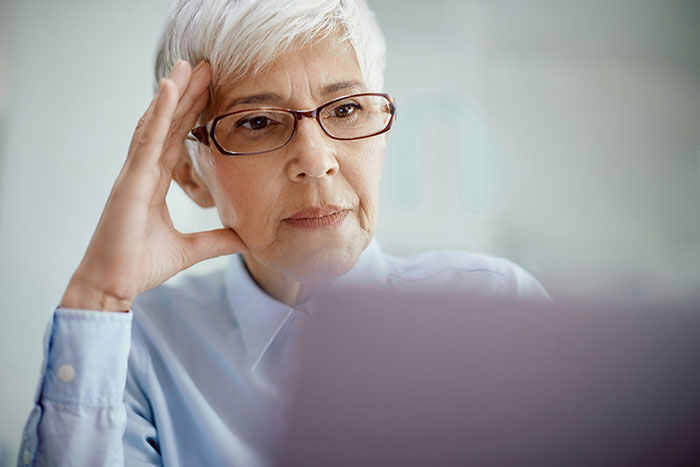 Older woman with glasses looking thoughtfully at a laptop, planning to hire a PI to uncover secrets about boyfriend