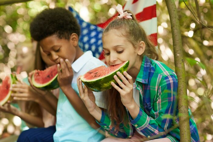 Children enjoying watermelon outdoors with an American flag in the background, illustrating parenting hacks for sanity.