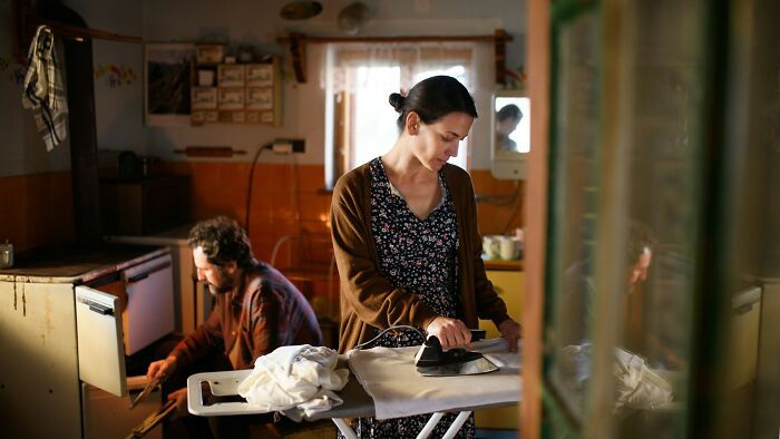 Woman ironing clothes and man checking oven in a worn kitchen, illustrating adults struggling with growing up responsibilities.