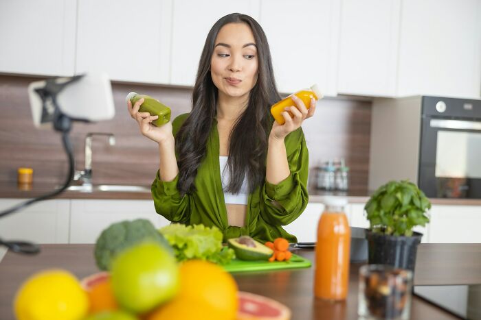 Young woman in modern kitchen holding green and orange juice bottles, showing a playful expression about traveling to the Bahamas.