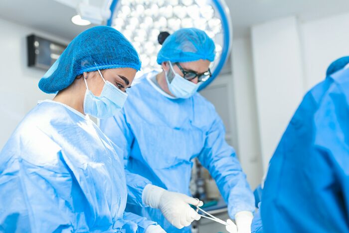 Two surgeons wearing blue scrubs and masks perform an operation under bright surgical lights in a medical setting.