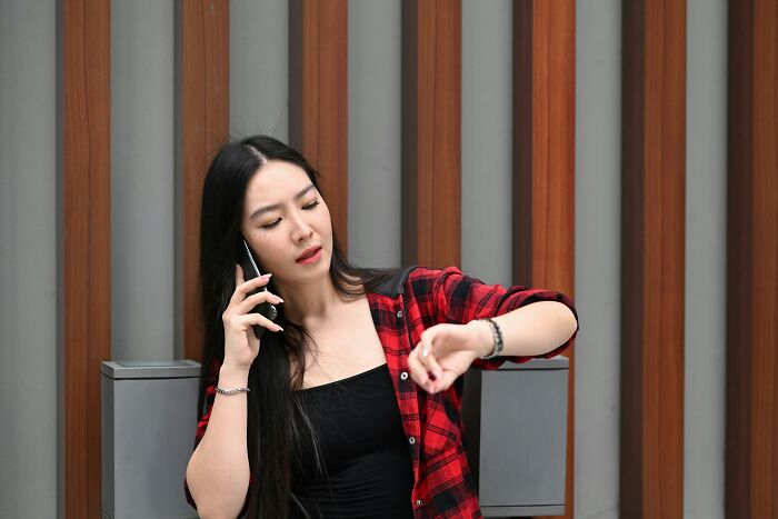 Woman in red plaid shirt talking on phone and checking watch against a wooden panel background, showing out-of-touch gestures.