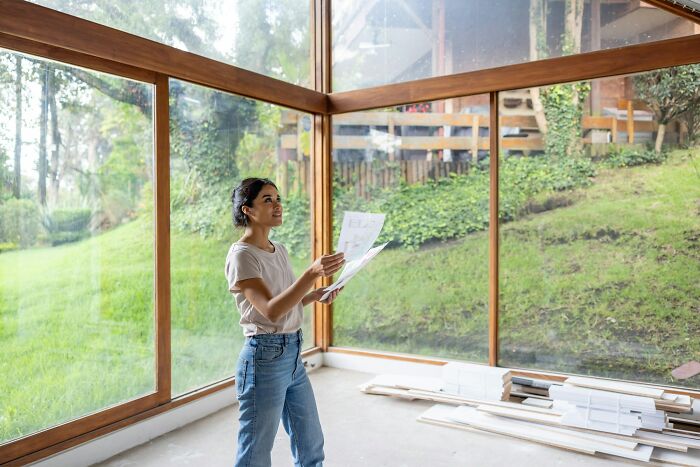Woman reviewing plans inside a spacious room with large windows overlooking a green outdoor area and construction materials.