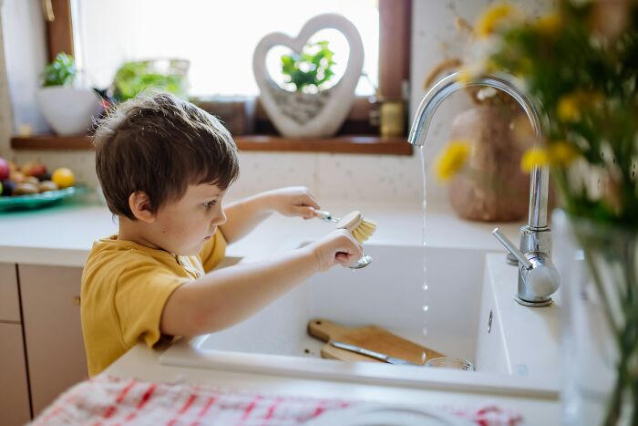 Young child using a brush to clean utensils in a kitchen sink, illustrating practical parenting hacks for saving sanity.