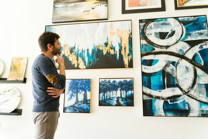 Man thoughtfully observing abstract and nature paintings in a gallery, reflecting on traveling to the Bahamas complaints.