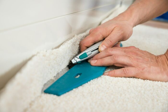 Hands using a cutting tool and a straightedge to trim carpet near the baseboard during a flooring installation.