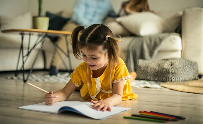 Young girl drawing with colored pencils on floor while parents relax on couch nearby, illustrating parenting hacks.
