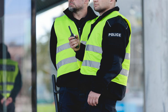 Two police officers in yellow safety vests standing outdoors, one holding a walkie-talkie during a security check.