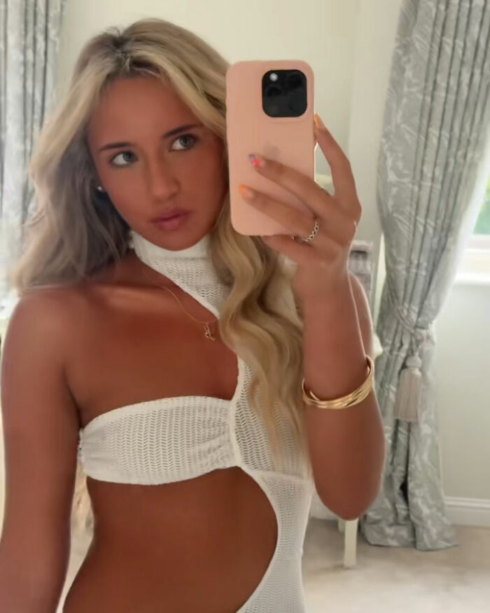 Glamorous law student taking a mirror selfie with long blonde hair, wearing a white cutout top indoors.