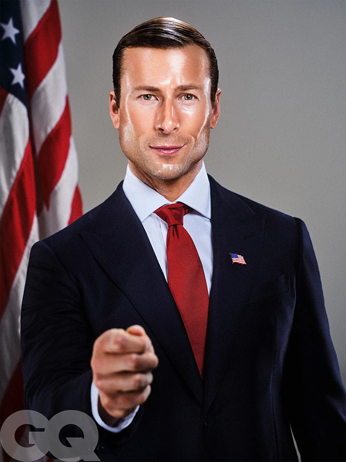 Glen Powell in a sharp suit and red tie posing confidently in front of an American flag during a GQ shoot. Glen Powell in a sharp suit and red tie posing confidently in front of an American flag during a GQ shoot.