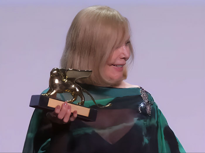 Kim Novak holding a Lifetime Achievement Award at Venice Film Festival, wearing a green dress and smiling.