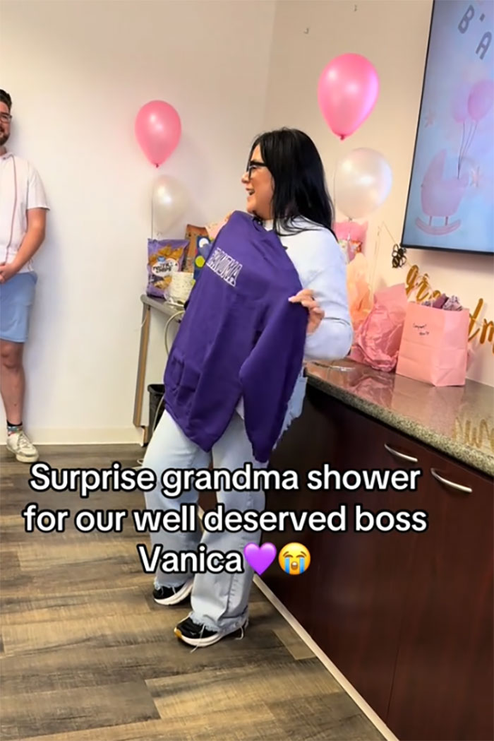 Woman at a surprise grandma baby shower holding a purple sweatshirt, celebrating her new status with balloons and gifts nearby.