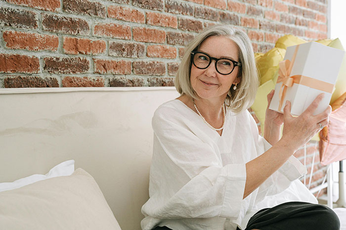 Smiling grandma in glasses holding a wrapped gift, celebrating with a baby shower for her new status.