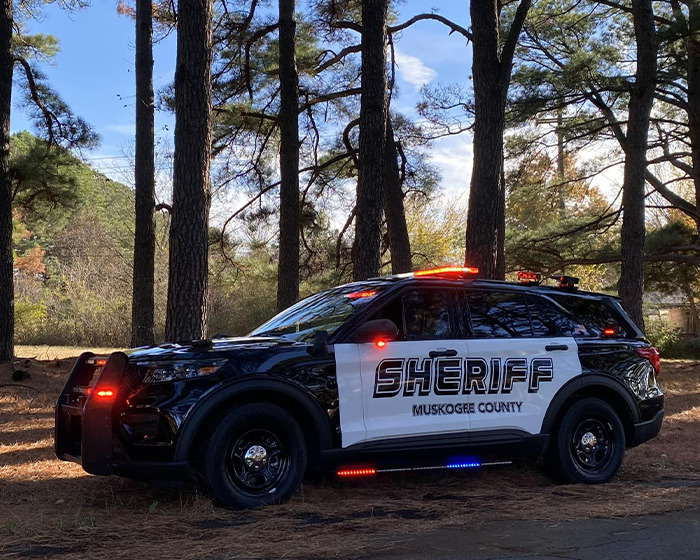 Muskogee County sheriff vehicle parked in a wooded area with lights flashing during an ongoing investigation.