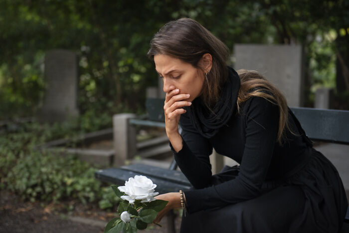 Woman sitting on a bench in a cemetery, holding a white rose, reflecting on things people would never admit in real life.