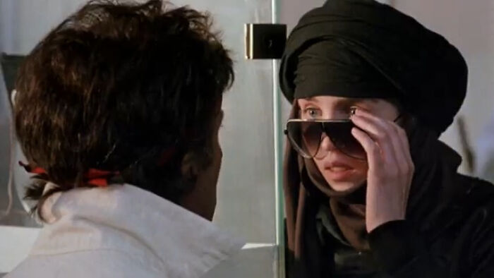 A scene from a worst-best movie showing a woman in dark clothing and sunglasses facing a man in a white jacket.