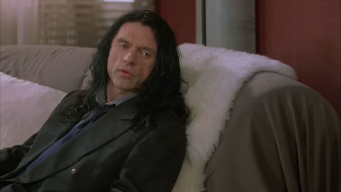 Scene from a worst-best movie showing a man with long black hair sitting on a couch, reflecting cult classic film style.