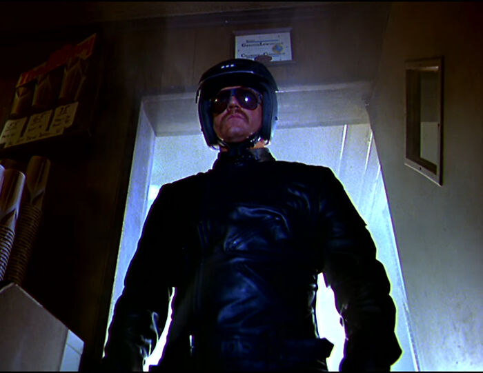 Man wearing black leather jacket, helmet, and sunglasses standing in a dimly lit room from worst-best movies collection.