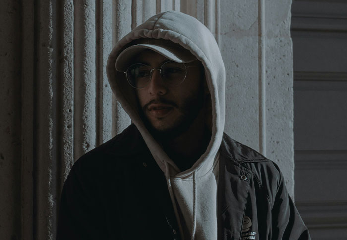 Man wearing glasses and a hooded jacket, standing against a textured wall, illustrating listening to instincts concept.