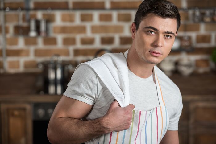 Young man in striped apron holding towel over shoulder in a rustic kitchen illustrating easy tricks to improve your kitchen game.