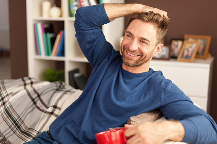 Smiling man in blue shirt relaxing on a couch with a mug, conveying a lighthearted schizophrenic man prank hospital visit moment.