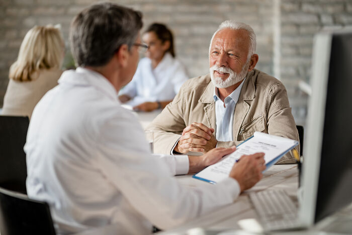 Senior man talking to doctor with a clipboard, illustrating advanced stupid out-of-touch comments in a clinical setting.