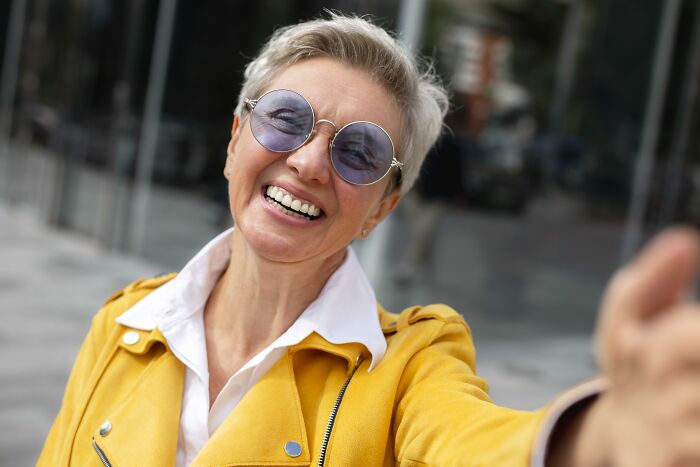 Smiling woman in yellow jacket and round sunglasses enjoying a bright day, representing doctors and nurses experiences.
