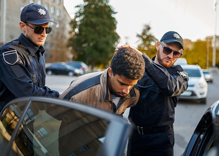 Man being arrested by two police officers outdoors near parked cars on a city street, woman considers canceling holiday booking.
