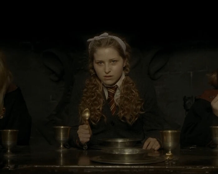 Harry Potter star seated at a table in a dark room wearing a school uniform with a Gryffindor tie.