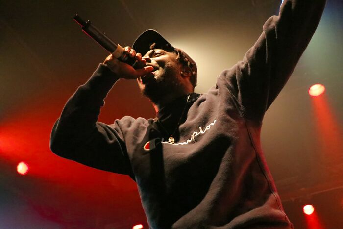 Performer wearing a black sweatshirt and cap singing passionately into a microphone on stage with warm lighting.