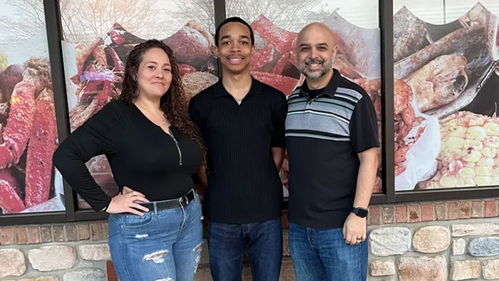 Three people standing together smiling outdoors with a stone wall and food images in the background, chilling messages theme.