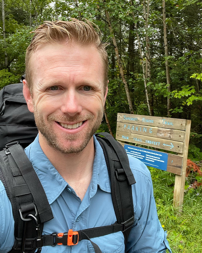 Smiling hiker wearing blue shirt and backpack near forest trail sign, related to hiker’s tragic last text and mountain incident. Smiling hiker wearing blue shirt and backpack near forest trail sign, related to hiker’s tragic last text and mountain incident.