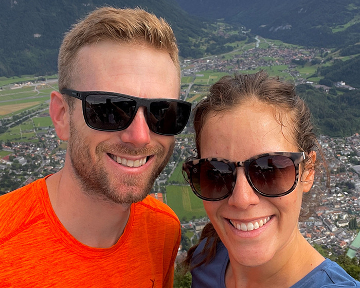 Young couple wearing sunglasses smiling during a mountain hike, with a village visible far below at high altitude Young couple wearing sunglasses smiling during a mountain hike, with a village visible far below at high altitude