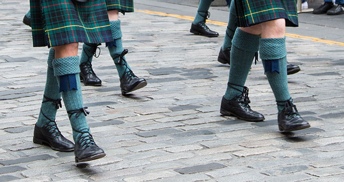 Men wearing traditional Scottish attire with tartan kilts and high socks walking on a cobblestone street in a historic bruha moment