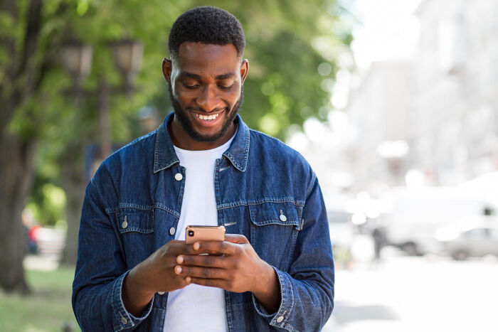 Smiling man in denim jacket texting on his phone outdoors, illustrating signs of the worst partner ever behavior.