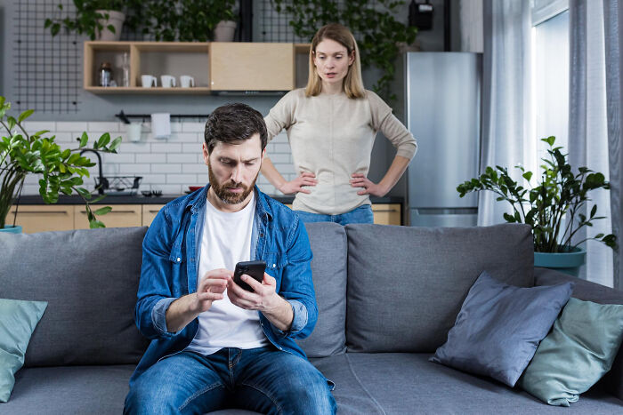 Couple arguing at home with man focused on phone and woman standing behind showing frustration, depicting worst partner behavior.