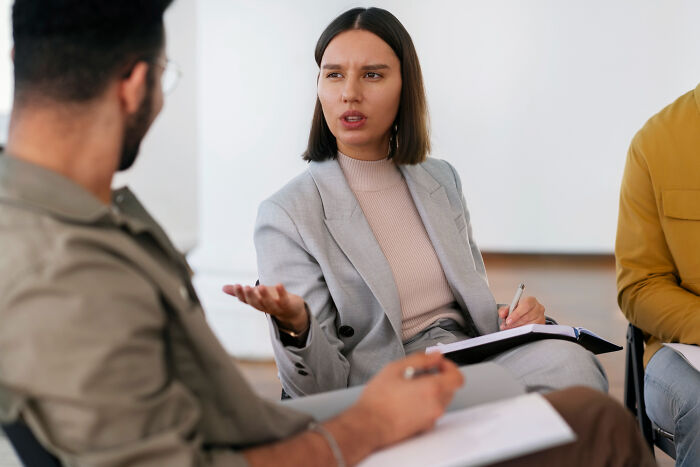 Woman in a gray suit explaining signs of a possible spouse during a serious conversation in a group setting.