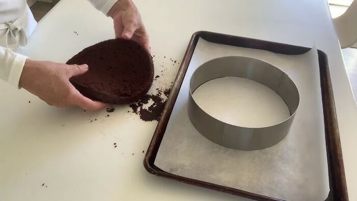 Hands holding a chocolate cake layer near a parchment-lined tray with a round baking mold, kitchen trick demonstration.